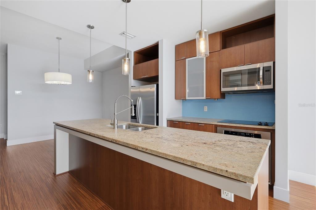 A kitchen features a granite-topped island, wood cabinetry, stainless steel appliances, and blue tile backsplash — 111 N 12TH STREET UNIT 1811, TAMPA, FL