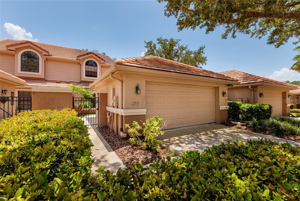 A two-story tan house features a single-car garage and a gated walkway with landscaping — 17557 FAIRMEADOW DRIVE, TAMPA, FL
