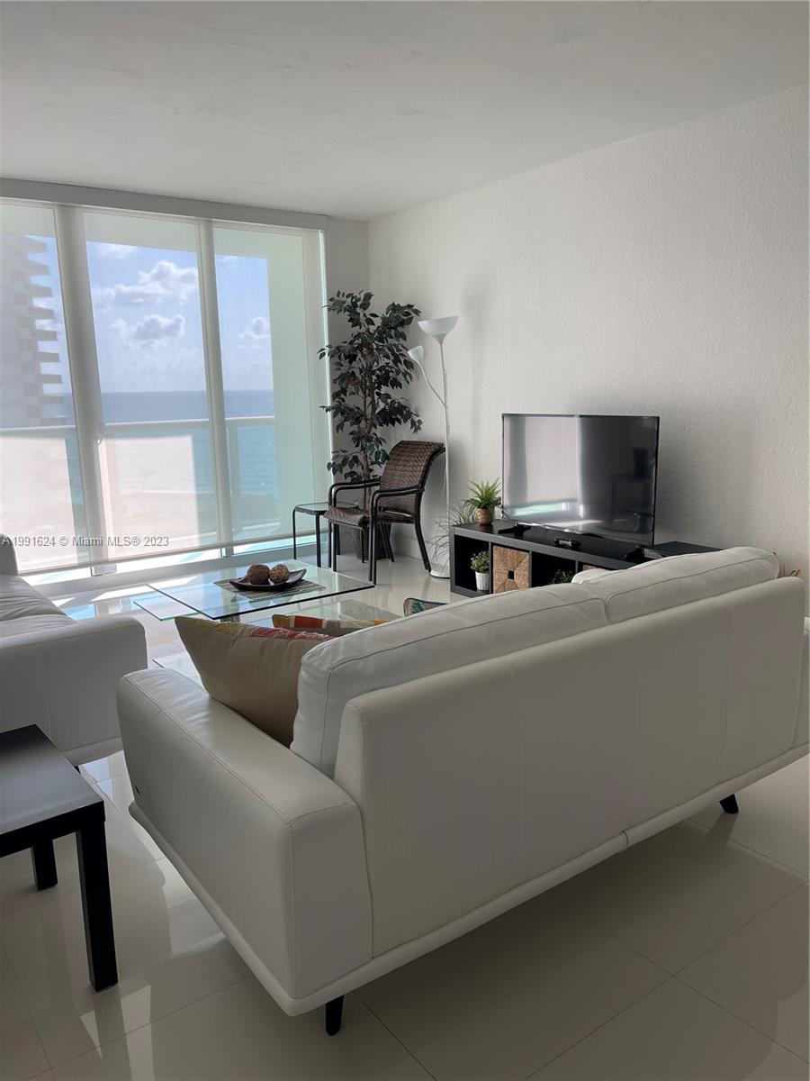 A living room features a white sofa, glass coffee table, television, and floor-to-ceiling windows — 3801 S OCEAN DR # 7X, Hollywood FL 33019, HOLLYWOOD, FL