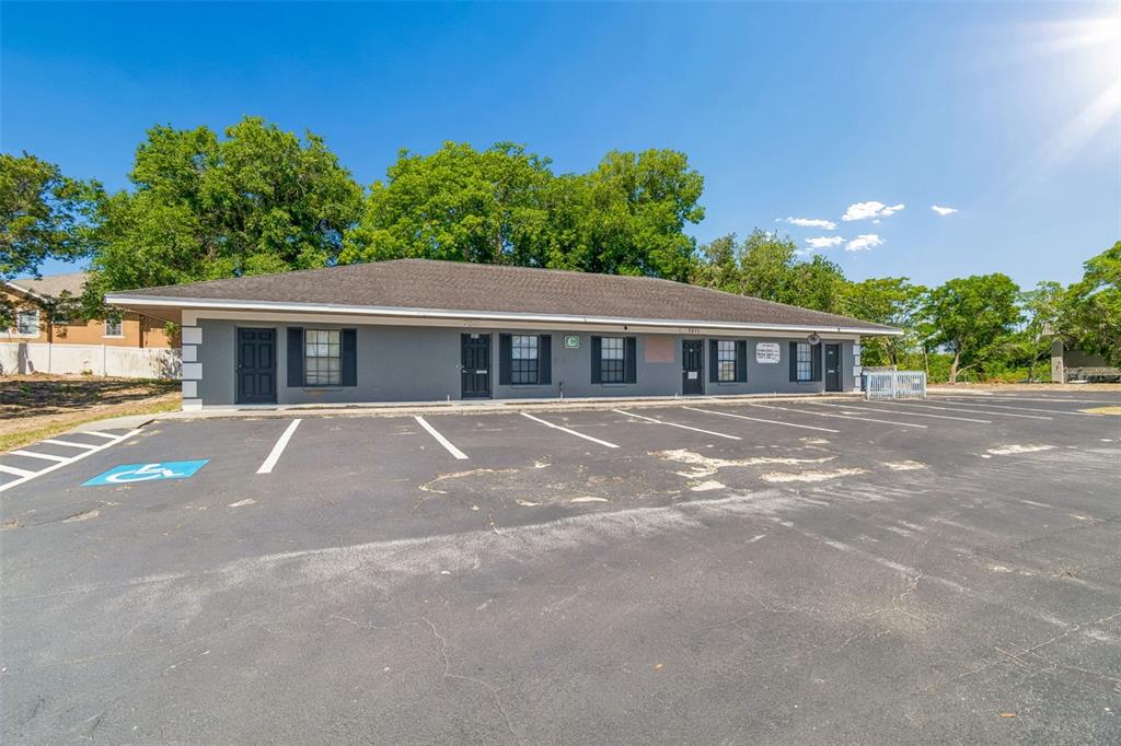 A single-story gray commercial building with multiple entrances sits behind a large, worn asphalt parking lot — 7511 LITTLE ROAD, NEW PORT RICHEY, FL