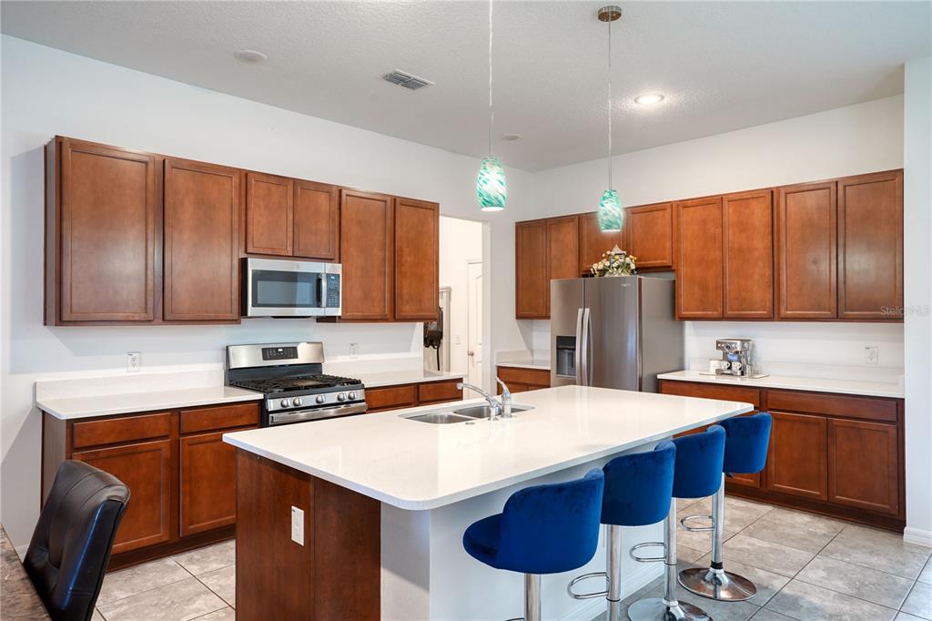 A kitchen features brown wooden cabinets, a white quartz island with blue barstools, and stainless appliances — 2052 EMERALD SPRINGS DRIVE, APOPKA, FL