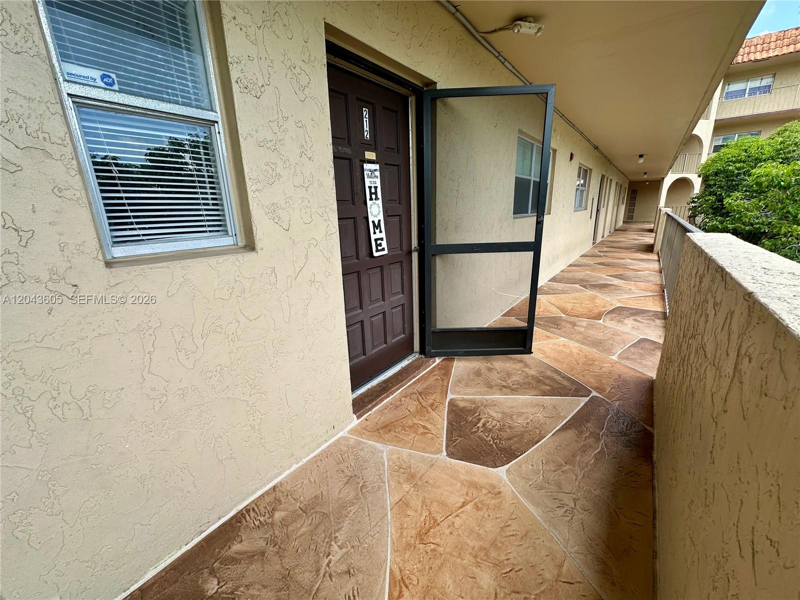 An exterior walkway with patterned concrete flooring leads to a brown door numbered 212 — 6201 N Falls Cir Dr # 212, Lauderhill FL 33319, LAUDERHILL, FL