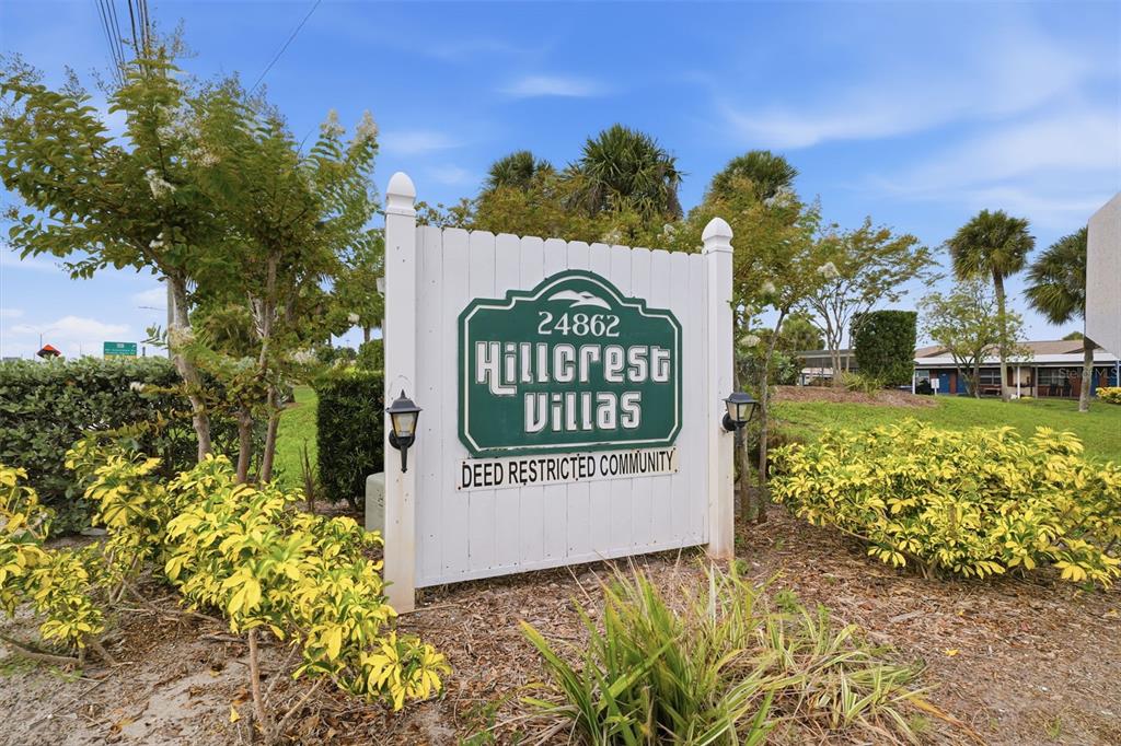 A white sign for Hillcrest Villas stands in a landscaped area with trees and shrubs — 24862 US HIGHWAY 19 N UNIT 1705, CLEARWATER, FL