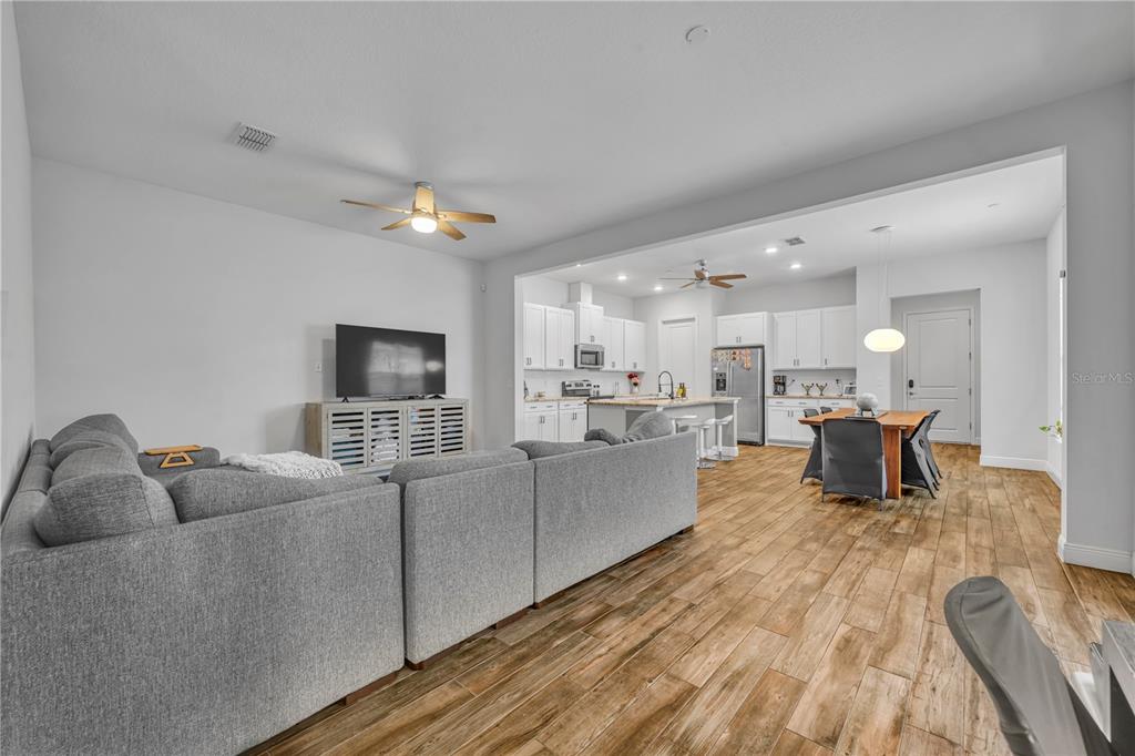 An open-concept living room and kitchen featuring gray sectional seating, wood-look tile flooring, and white cabinetry — 13433 STODDART AVENUE, ORLANDO, FL