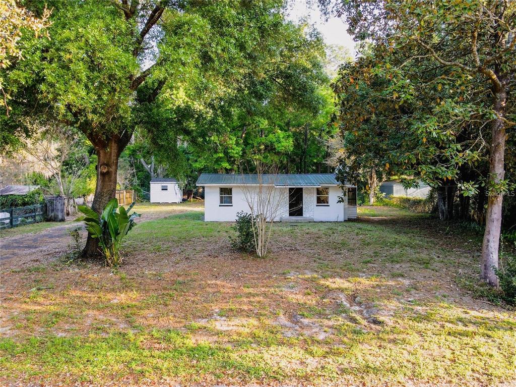 A small, white, single-story house sits on a grassy lot surrounded by mature trees — 107 EARLS LANE, APOPKA, FL