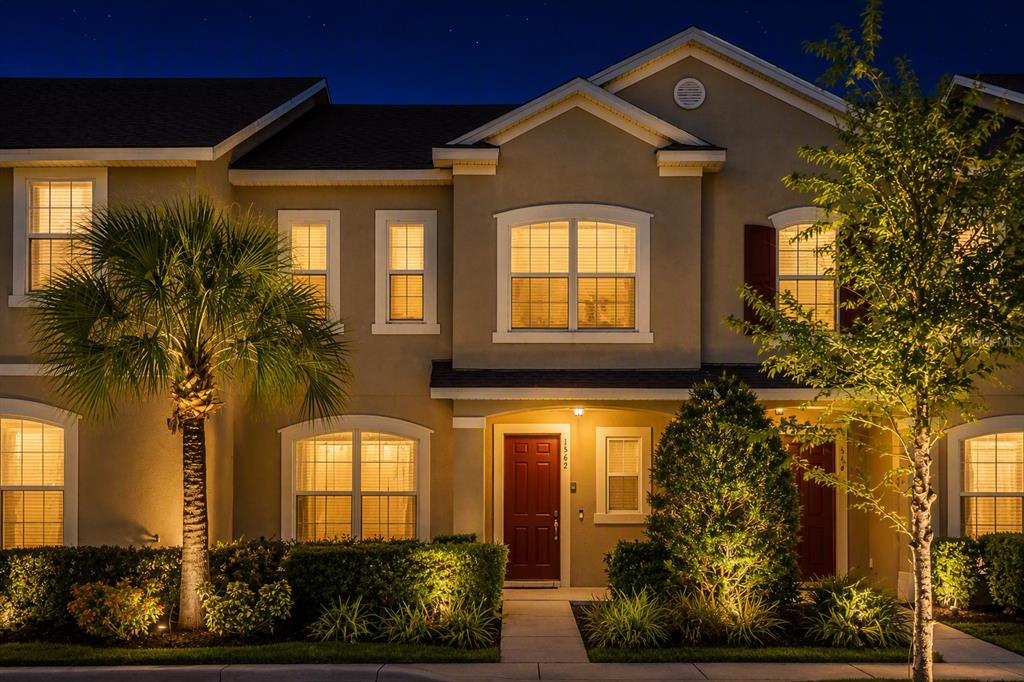 A two-story townhouse exterior at dusk with a red front door and illuminated windows — 1562 CAREY PALM CIRCLE, KISSIMMEE, FL