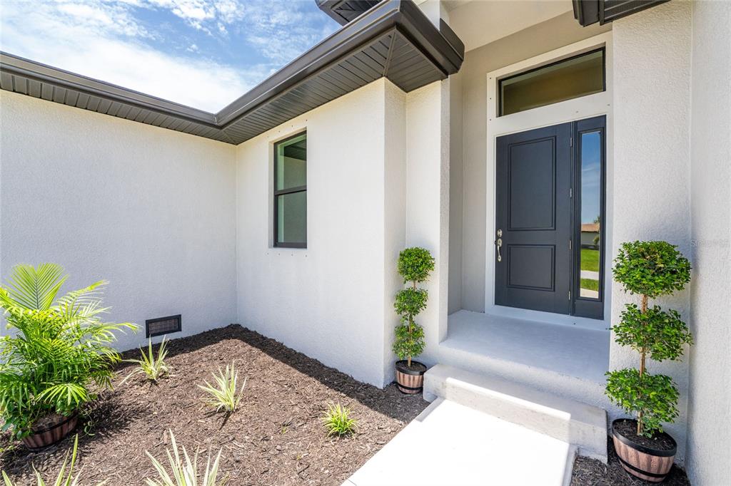 A modern home entrance features a dark gray door, white stucco walls, and potted topiaries — 9590 HONEYMOON DRIVE, PORT CHARLOTTE, FL