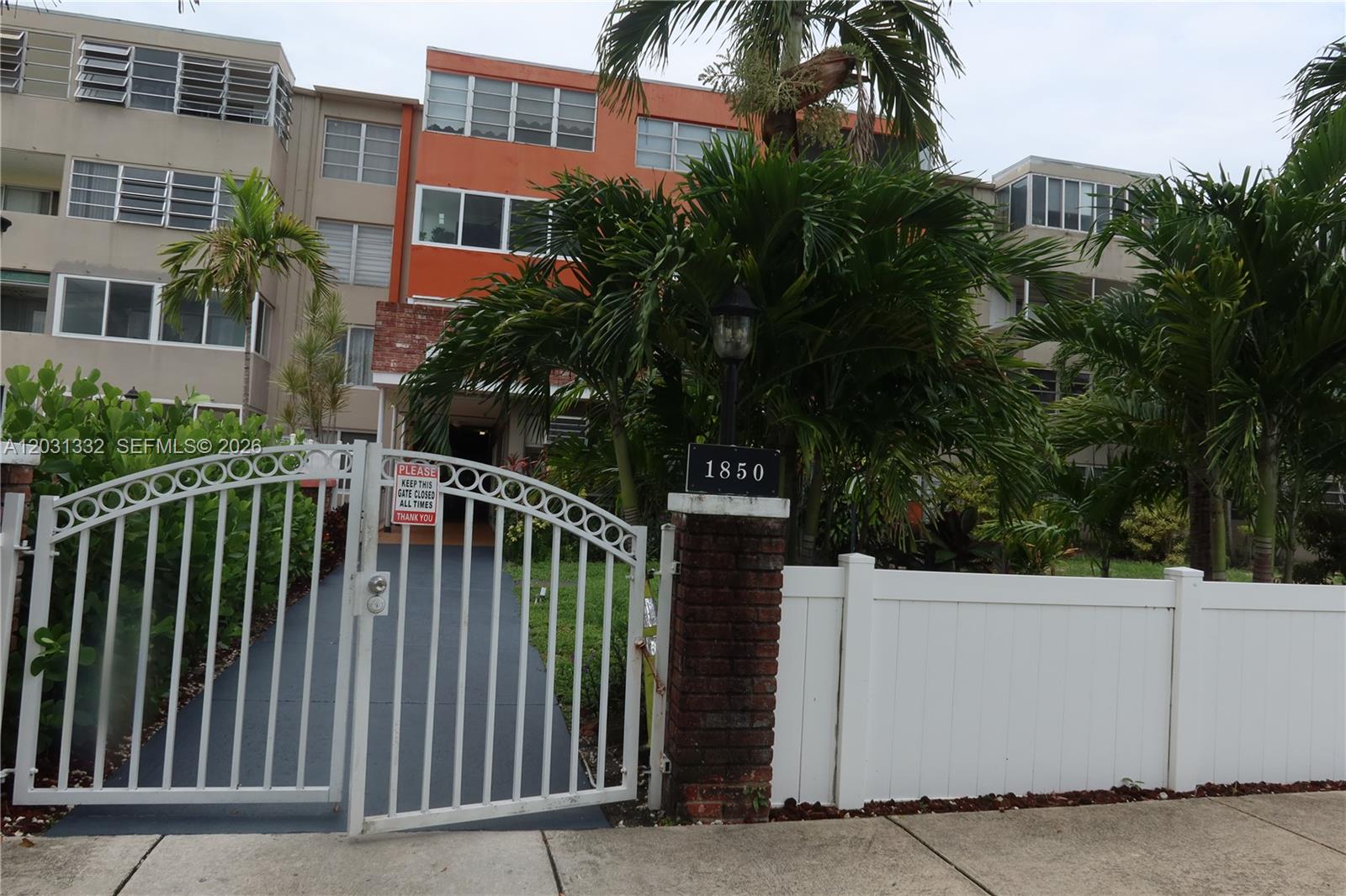 A white metal gate stands before a multi-story apartment building with orange accents — 1850 NE 169th St # 312, North Miami Beach FL 33162, NORTH MIAMI BEACH, FL