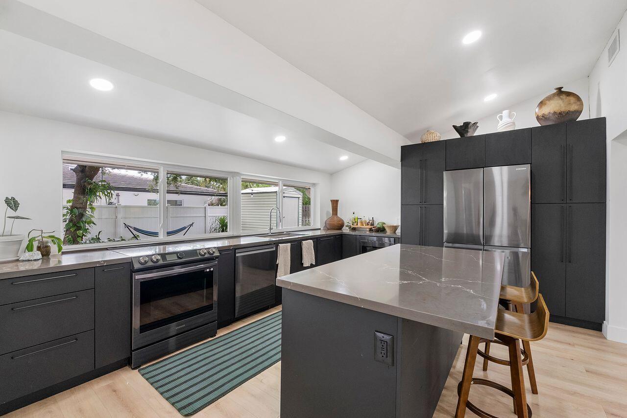 A modern kitchen features dark cabinetry, a quartz island, stainless steel appliances, and large windows — 701 SE 5th Court, Pompano Beach FL 33060, POMPANO BEACH, FL