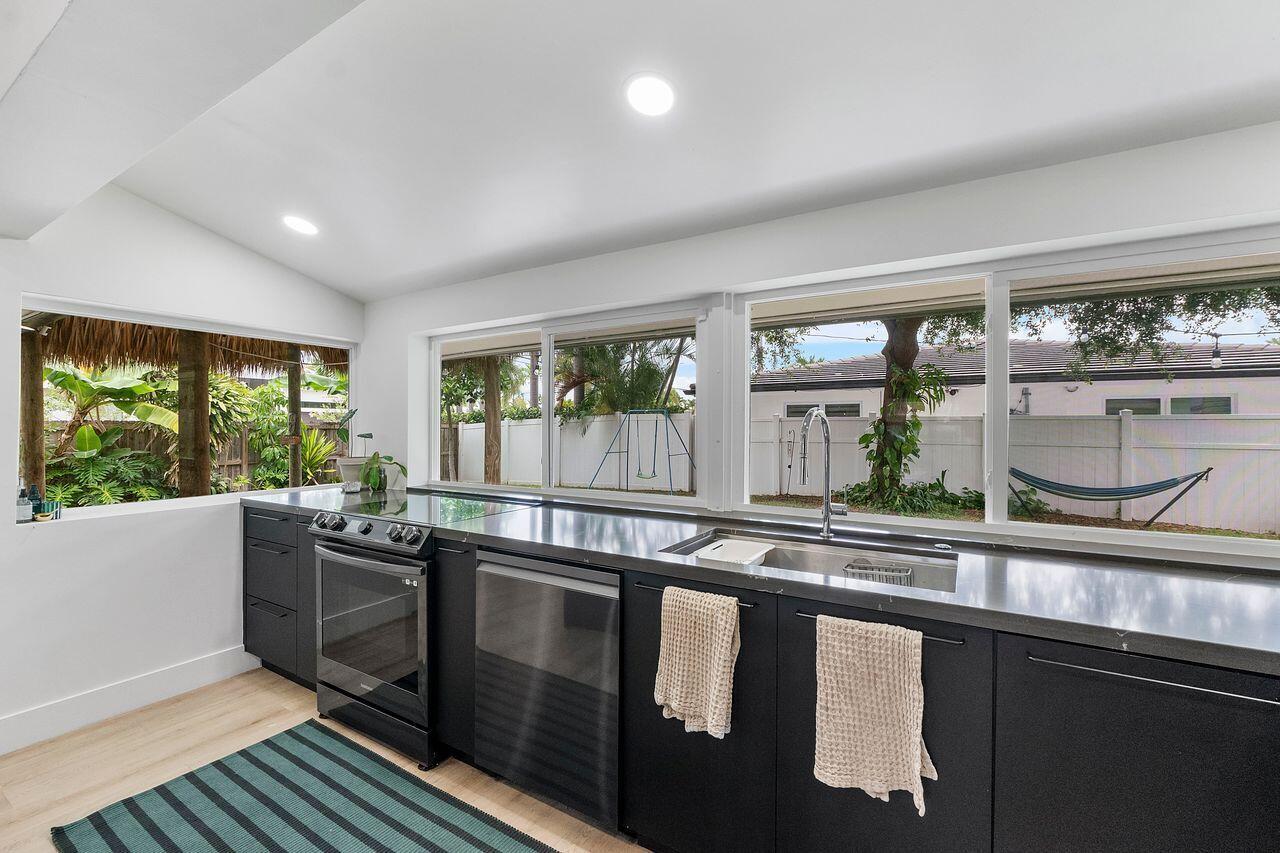 A modern kitchen features black cabinets, dark countertops, and large windows overlooking a backyard — 701 SE 5th Court, Pompano Beach FL 33060, POMPANO BEACH, FL