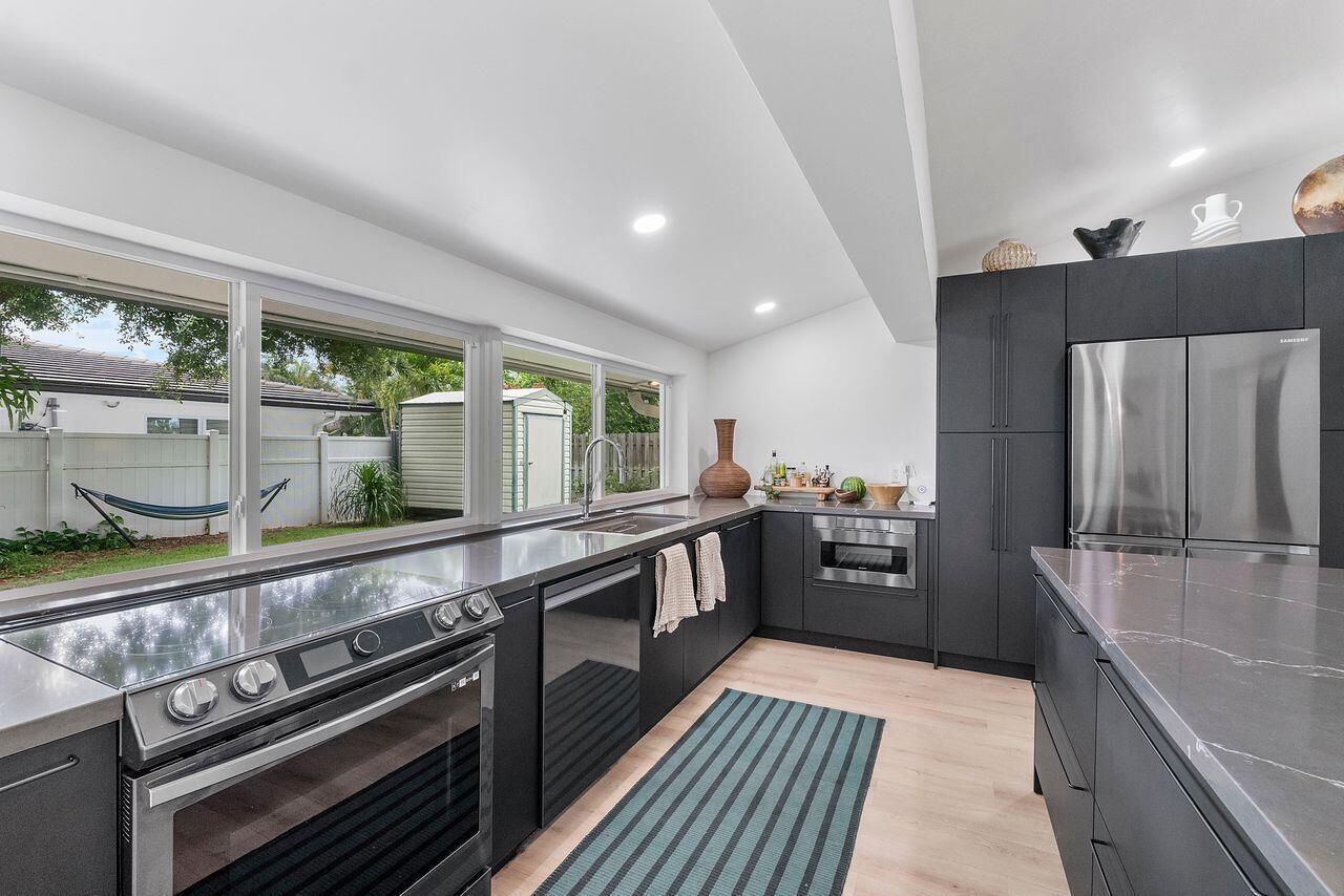 A modern kitchen features dark cabinetry, stainless steel appliances, quartz countertops, and large windows overlooking a yard — 701 SE 5th Court, Pompano Beach FL 33060, POMPANO BEACH, FL