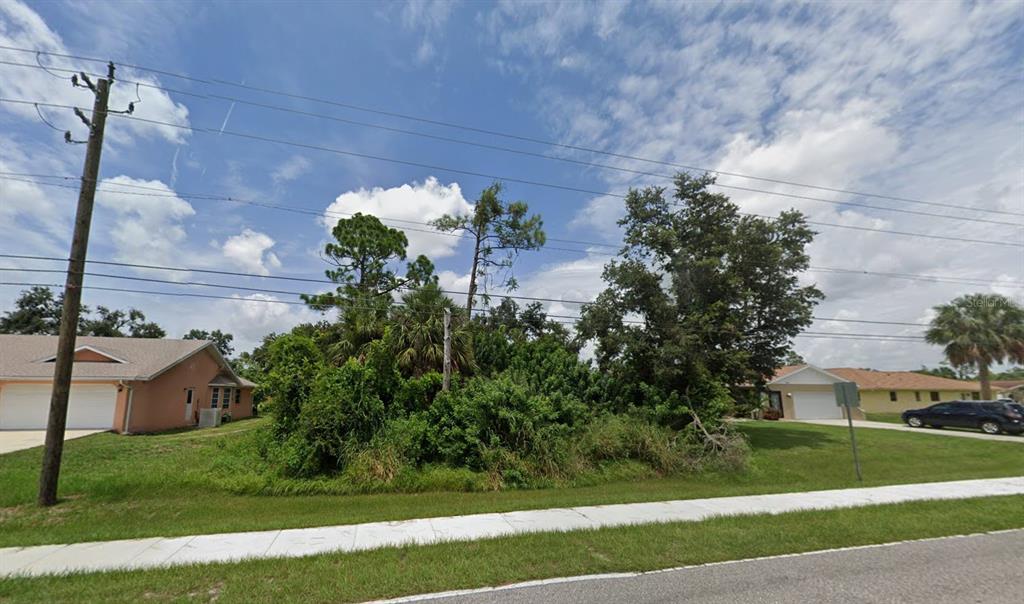 A vacant, overgrown lot sits between two suburban houses under a cloudy blue sky — 21086 PEACHLAND BOULEVARD, PORT CHARLOTTE, FL