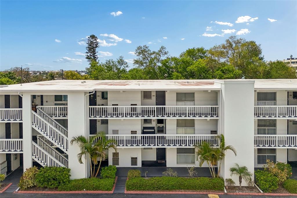 An exterior view of a three-story white apartment building with balconies and palm trees — 2944 W BAY DRIVE UNIT 106, BELLEAIR BLUFFS, FL