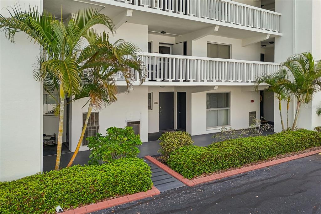 A two-story apartment building exterior features white walls, balconies, palm trees, and landscaped hedges — 2944 W BAY DRIVE UNIT 106, BELLEAIR BLUFFS, FL