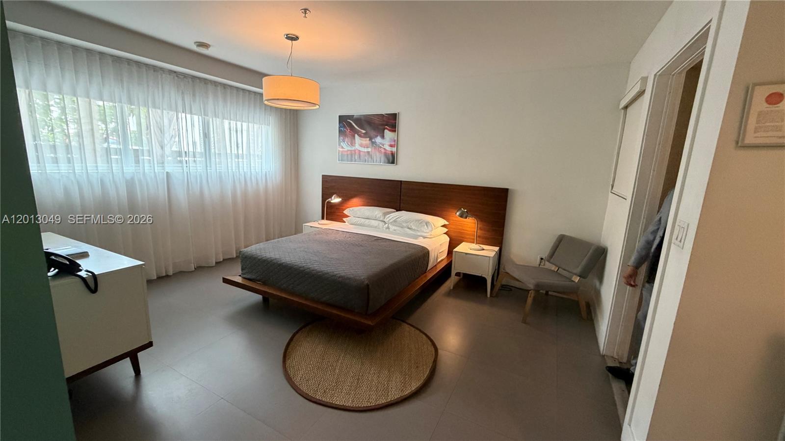 A bedroom features a platform bed with a wood headboard, gray tile floors, and sheer curtains — 1816 Meridian Ave # 9, Miami Beach FL 33139, MIAMI BEACH, FL