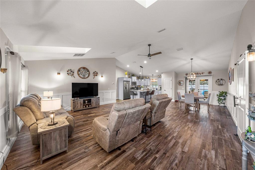 An open-concept living room and kitchen featuring vaulted ceilings, wood-look flooring, and white wainscoting — 6658 SW 109TH LANE, OCALA, FL