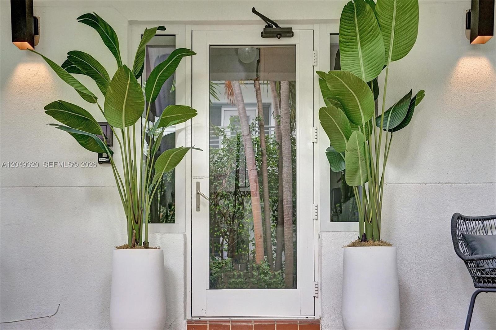 A white door is flanked by two large potted plants and wall-mounted outdoor lights — 1431 Lincoln Ter # 3, Miami Beach FL 33139, MIAMI BEACH, FL