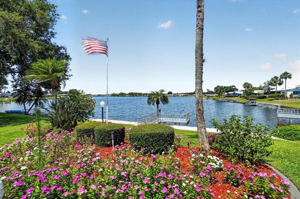 A waterfront view features a manicured garden with pink flowers, a flag, and a dock — 8469 IMPERIAL LAKES CIRCLE, PALMETTO, FL