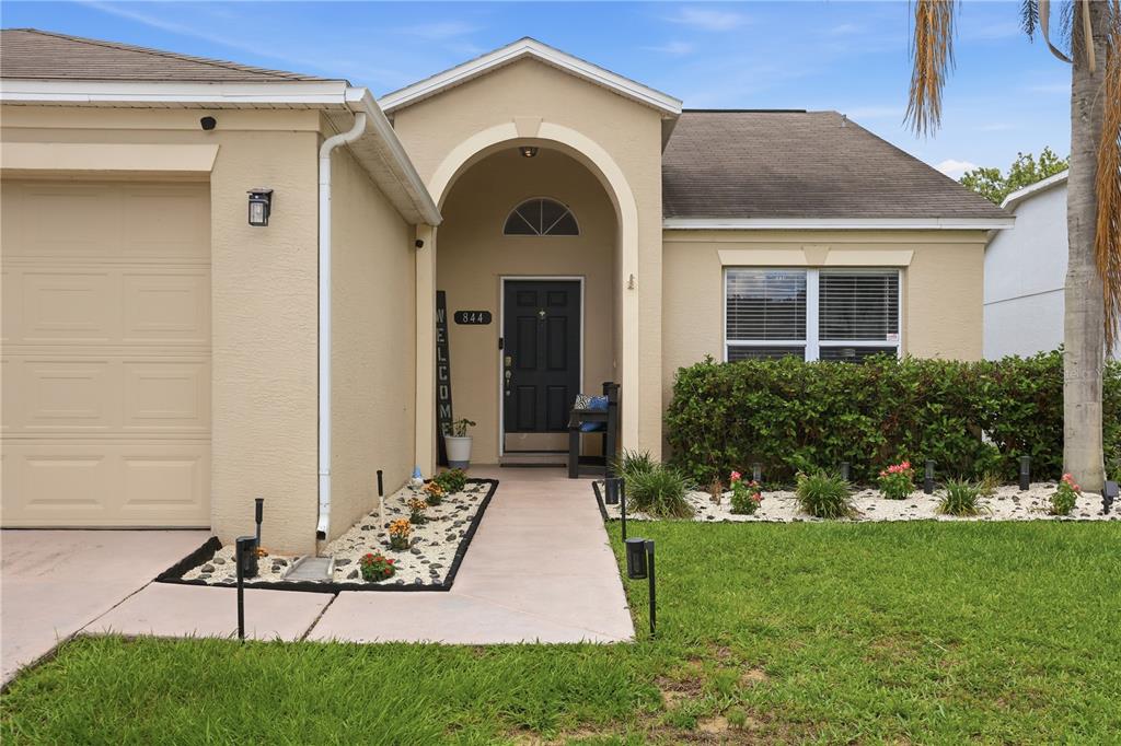 A single-story house exterior features a tan stucco facade, a garage, and a covered entryway — 844 WOODSONG WAY, CLERMONT, FL