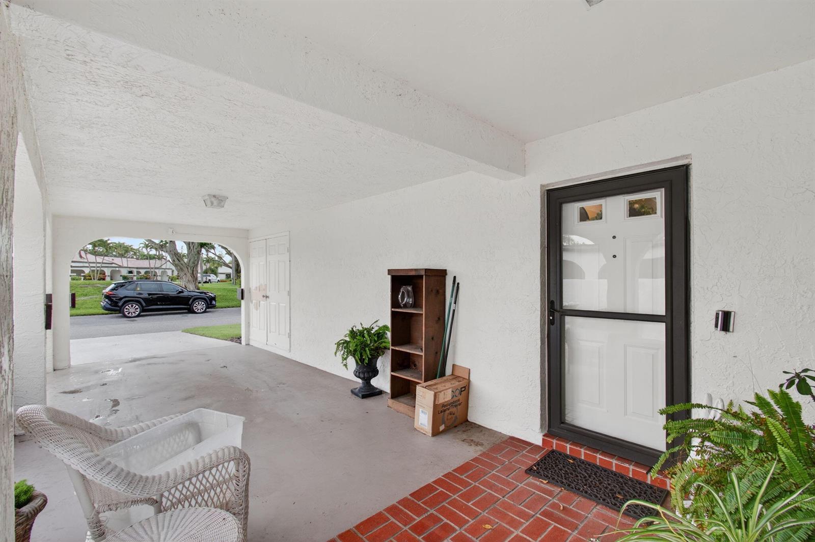 A covered carport area features a white front door, brick flooring, and a wicker chair — 5707 Parkwalk Circle E, Boynton Beach FL 33472, BOYNTON BEACH, FL