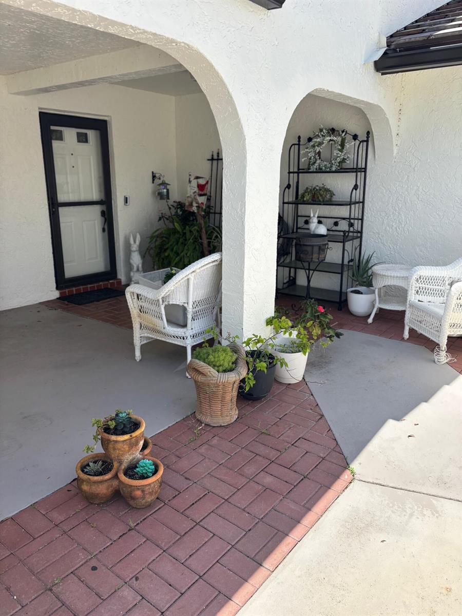 A covered porch features white wicker chairs, potted plants, and a black metal shelving unit — 5707 Parkwalk Circle E, Boynton Beach FL 33472, BOYNTON BEACH, FL