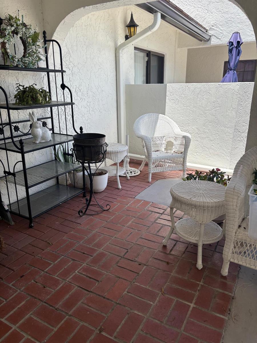A covered patio features red brick flooring, white wicker furniture, and a black metal shelf — 5707 Parkwalk Circle E, Boynton Beach FL 33472, BOYNTON BEACH, FL