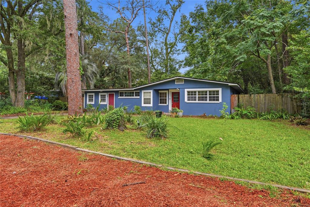 A blue single-story house with a red front door sits on a grassy, wooded lot — 630 NW 35TH TERRACE, GAINESVILLE, FL