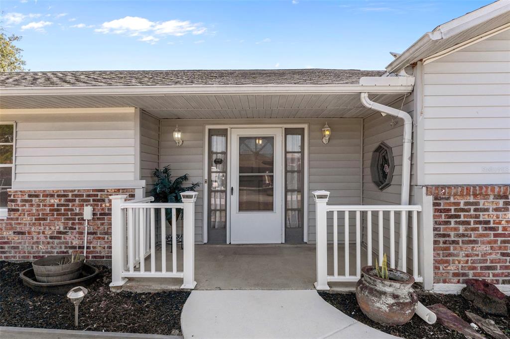 A front porch with a white door, sidelights, and railings sits between brick exterior walls — 4286 BLUEWATER AVENUE, SPRING HILL, FL