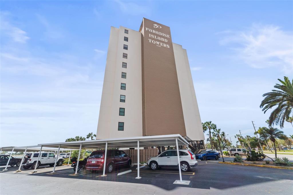 A tall, multi-story apartment building stands behind a parking lot with covered carports — 10355 PARADISE BOULEVARD UNIT 403, TREASURE ISLAND, FL