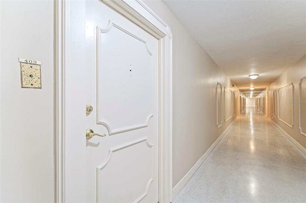 A white door with gold hardware leads into a long, brightly lit apartment building hallway — 10355 PARADISE BOULEVARD UNIT 403, TREASURE ISLAND, FL