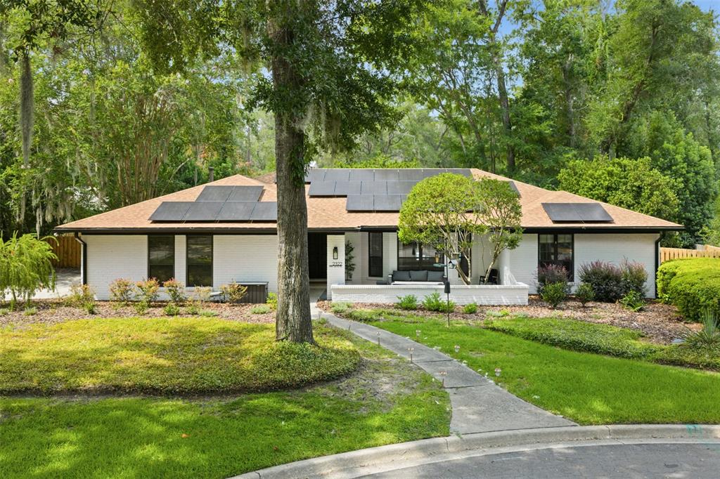 A single-story white brick house with a shingled roof and solar panels sits behind trees — 2322 NW 14TH PLACE, GAINESVILLE, FL