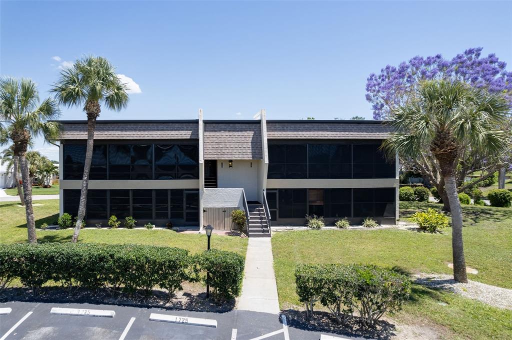 A two-story building with dark screened-in balconies and a central staircase sits behind a parking lot — 1727 BONITAS CIRCLES UNIT 1718-C, VENICE, FL