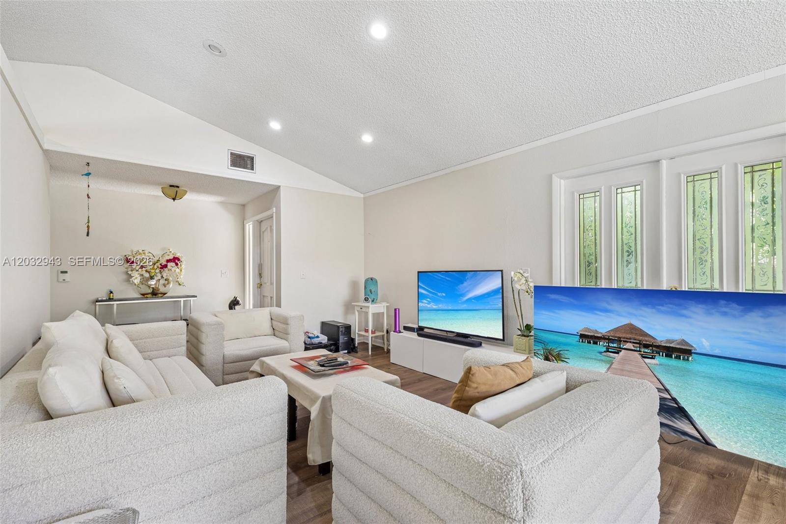 A living room features white textured furniture, a vaulted ceiling, and a large television screen — 7160 NW 4th Ave, Boca Raton FL 33487, BOCA RATON, FL