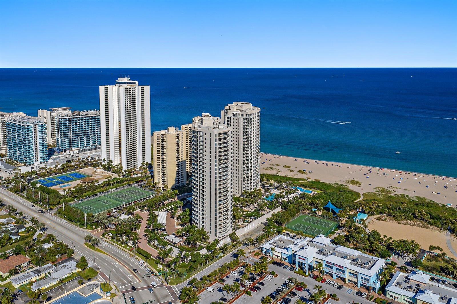 An aerial view shows high-rise residential buildings, tennis courts, and a beach along the ocean — 2700 N Ocean Drive # 1b, Riviera Beach FL 33404, RIVIERA BEACH, FL
