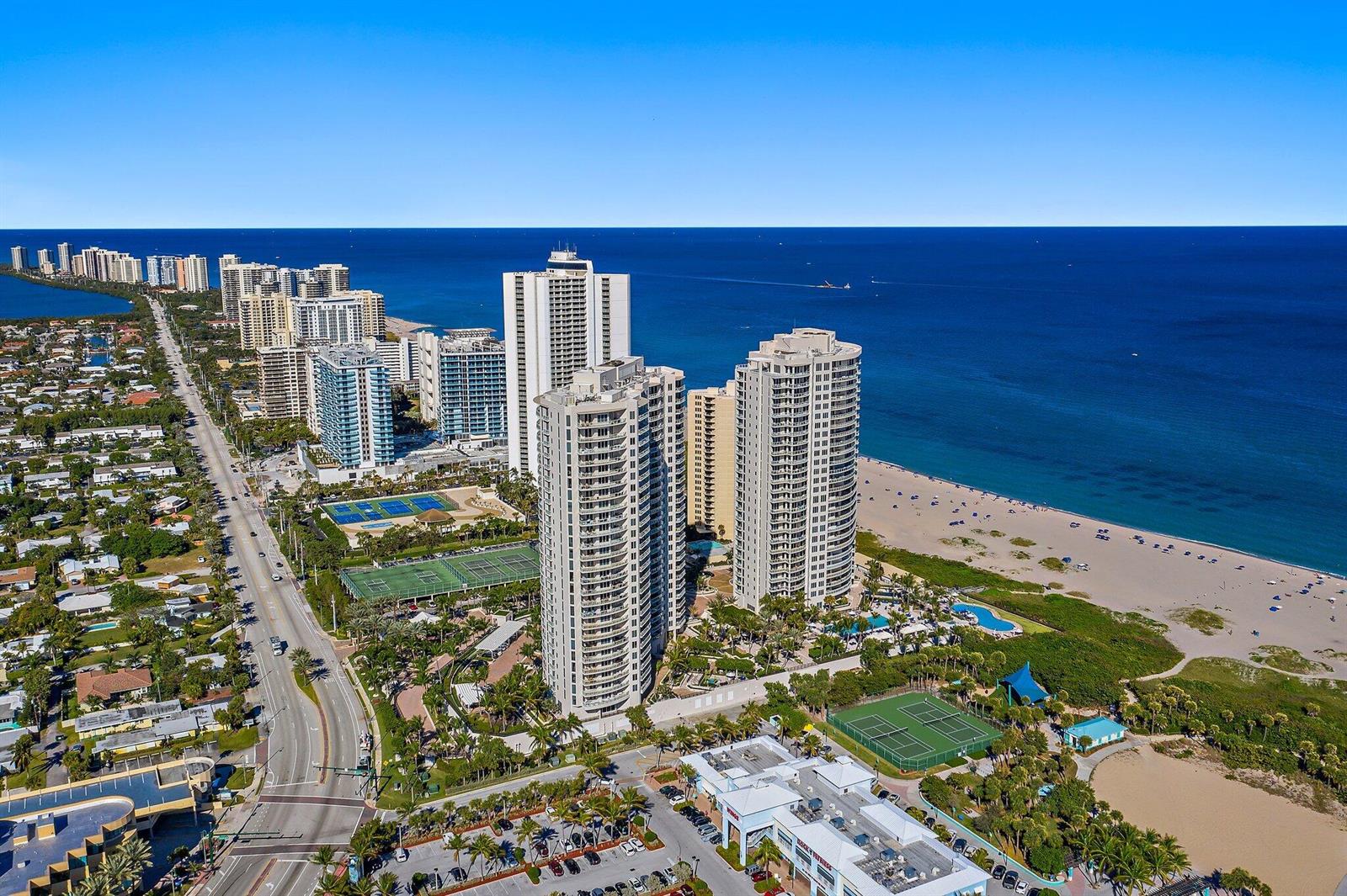 An aerial view shows high-rise residential towers along a beach with tennis courts and roads — 2700 N Ocean Drive # 1b, Riviera Beach FL 33404, RIVIERA BEACH, FL
