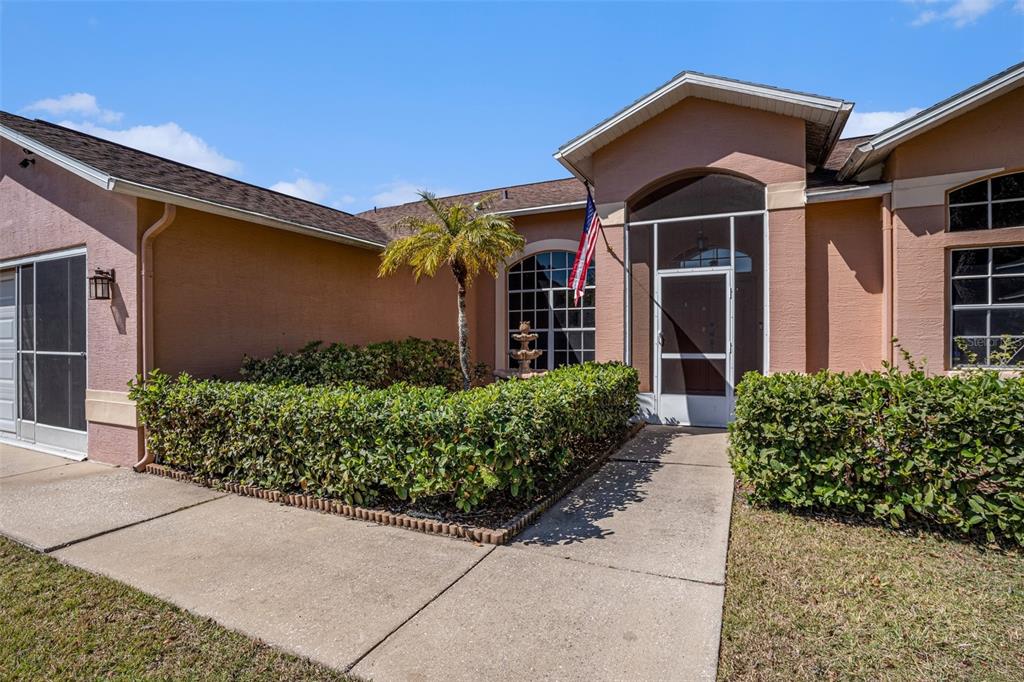 The exterior of a tan house features a screened entryway, a small palm tree, and landscaping — 7741 DAMASK LANE, NEW PORT RICHEY, FL