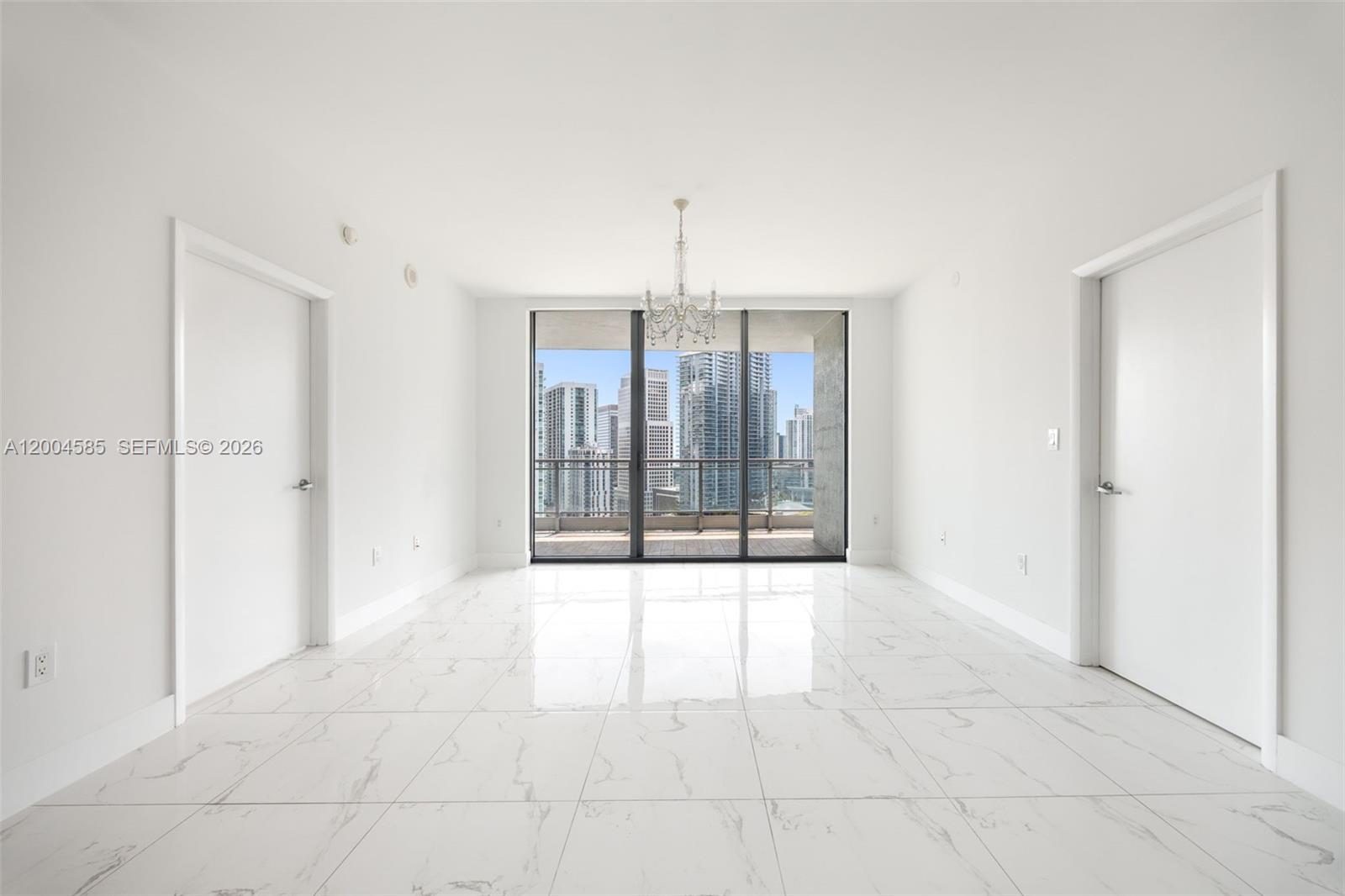 A bright, empty room features white marble-look tile flooring, a central chandelier, and balcony access — 90 SW 3rd St # 2602, Miami FL 33130, MIAMI, FL