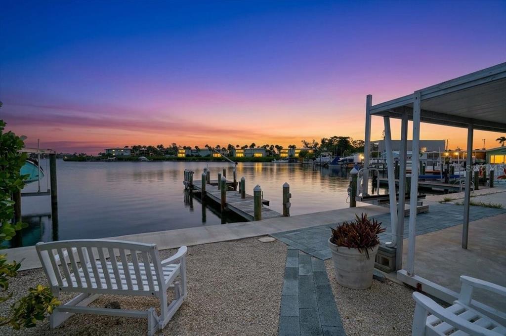 A white wooden bench sits on a gravel patio overlooking a boat dock at sunset — 17 SEASIDE COURT, HOLMES BEACH, FL
