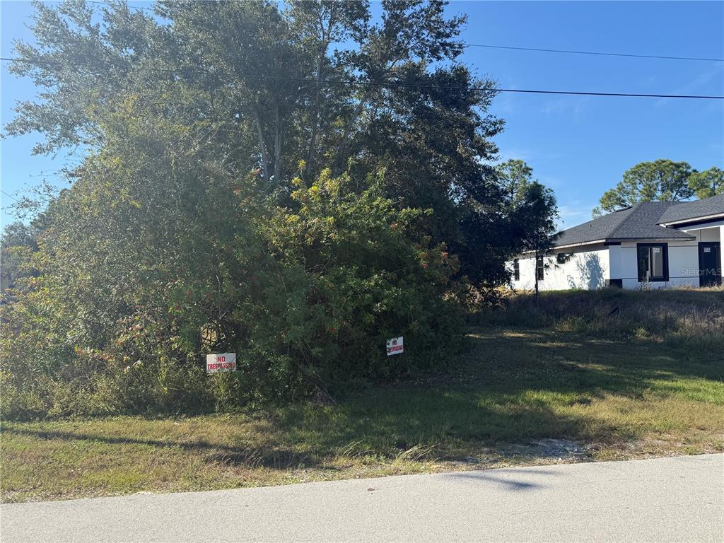 A vacant, tree-covered lot sits next to a newly constructed house under a blue sky — 205 GILBERT AVENUE N, LEHIGH ACRES, FL