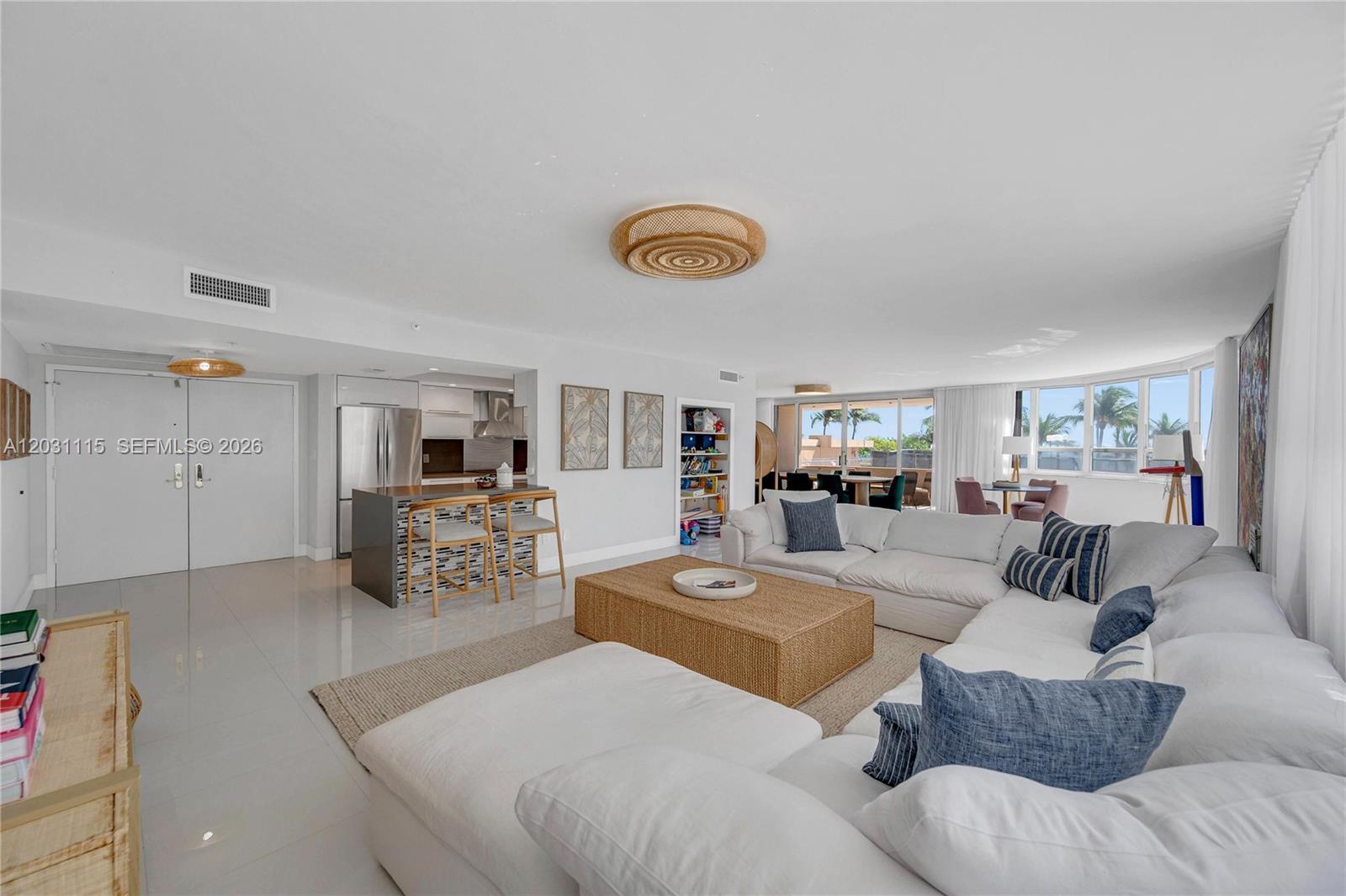 A bright, open-concept living room features a large white sectional, wicker coffee table, and kitchen — 10175 Collins Ave # 102, Bal Harbour FL 33154, BAL HARBOUR, FL