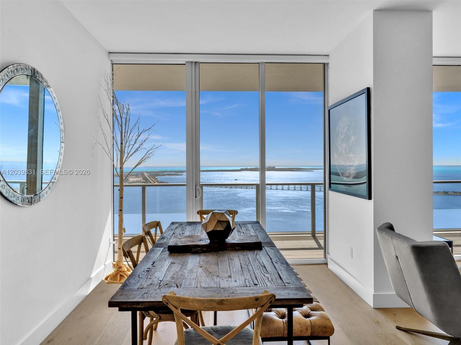 A dining room features a rustic wooden table, light wood floors, and floor-to-ceiling water views — 1300 E Brickell Bay Dr # 4202, Miami FL 33131, MIAMI, FL