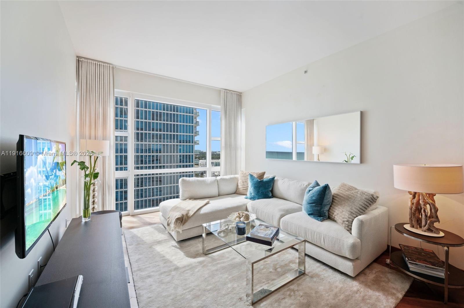 A modern living room features a white sectional sofa, glass coffee table, and floor-to-ceiling windows — 6801 Collins Ave, Miami Beach FL 33141, MIAMI BEACH, FL