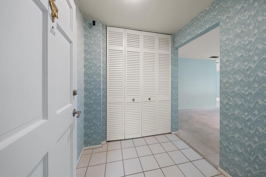 An entryway with white tiled floors, blue patterned wallpaper, and white louvered closet doors — 100 BLUFF VIEW DRIVE UNIT 114A, BELLEAIR BLUFFS, FL
