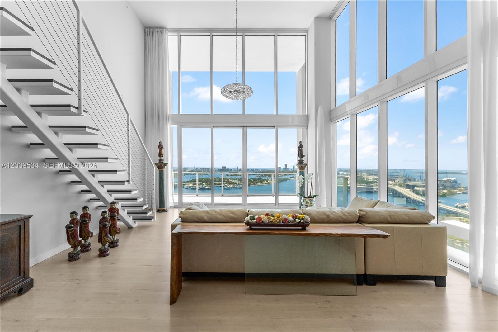 A modern living room features a floating staircase, floor-to-ceiling windows, and panoramic water views — 1040 Biscayne Blvd # 3101, Miami FL 33132, MIAMI, FL