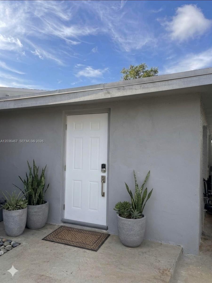 A gray exterior wall features a white front door with a smart lock and potted plants — 1321 NE 212th Ter # 2, Miami FL 33179, MIAMI, FL