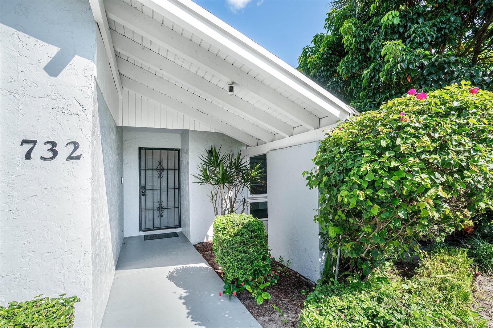 A white house exterior features a covered entryway with a metal security door and landscaping — 732 Lago Road, Delray Beach FL 33445, DELRAY BEACH, FL