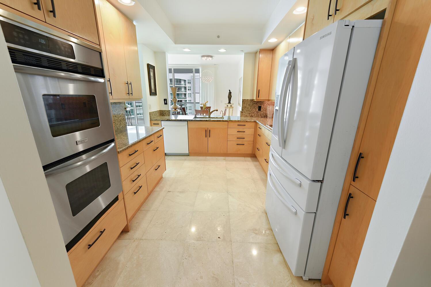A galley kitchen features light wood cabinets, stainless steel appliances, and beige tile flooring — 550 SE Mizner Boulevard # B702, Boca Raton FL 33432, BOCA RATON, FL