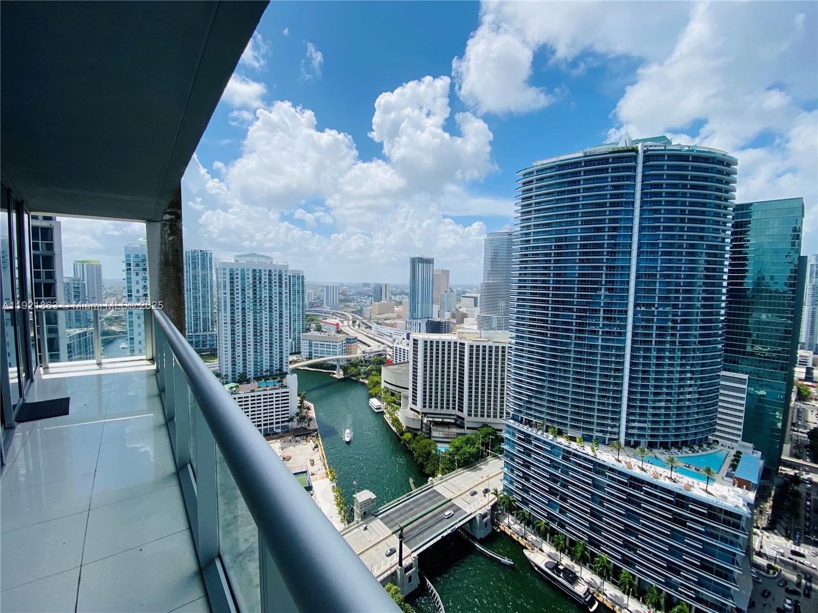 A high-rise balcony overlooks a river, a bridge, and surrounding city skyscrapers under a cloudy sky — 475 Brickell Ave # 3715, Miami FL 33131, MIAMI, FL
