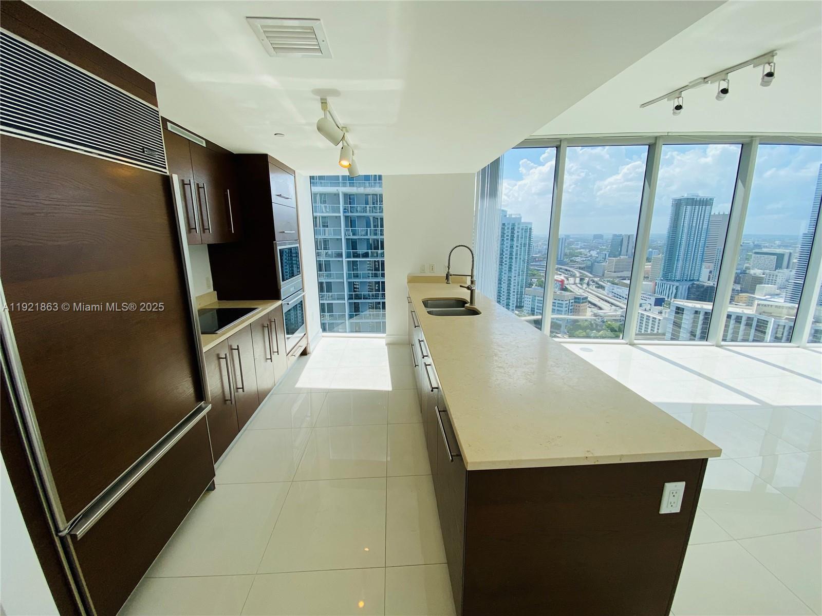 A modern kitchen features dark wood cabinetry, light countertops, and floor-to-ceiling windows with city views — 475 Brickell Ave # 3715, Miami FL 33131, MIAMI, FL