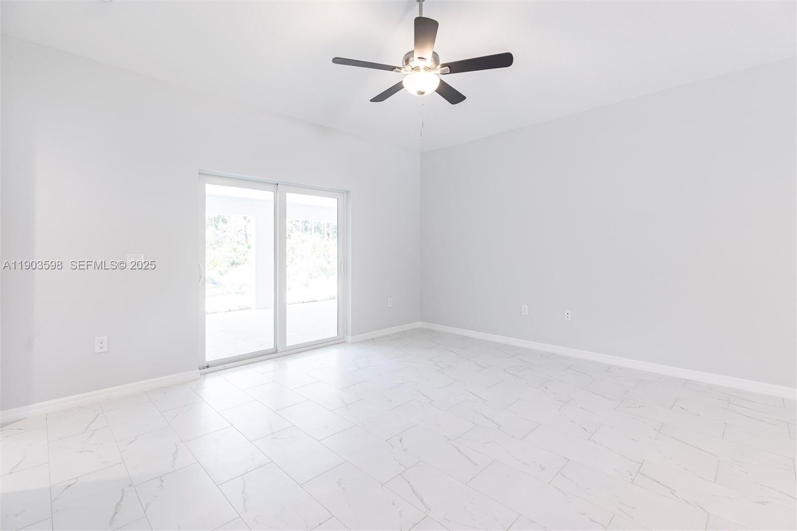 A bright, empty room features white marble-look tile flooring, a ceiling fan, and sliding doors — 2005 Rosa Ct, Lehigh Acres FL 33972, LEHIGH ACRES, FL